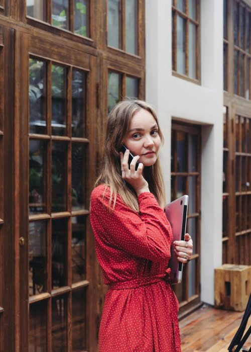 Caucasian woman in red polka dot dress talking on smartphone indoors, holding laptop.