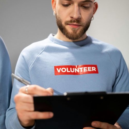 pexels-photo-6591150-6591150 Young adult male volunteer with beard writing on clipboard, wearing a volunteer shirt indoors.